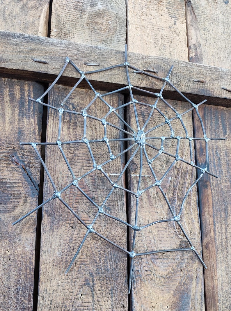 May include: A spider web sculpture crafted from dark grey metal rods. The design features a central hub with radiating spokes connected by concentric circular segments, all joined together with visible welded joints at each intersection point.