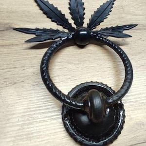 以下が含まれることがあります： 葉のデザインが施された黒い金属製のドアノッカー。ノッカーは中央にリングのある円形をしています。葉はリングの周りに星形に配置されています。