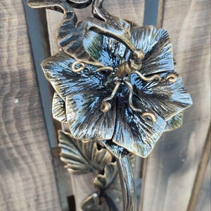 May include: A metal wall sculpture featuring a large, multi-petaled flower with a dark finish and gold-toned highlights. A stylised bird is positioned above the flower, and the piece includes detailed leaves on a curved stem, all crafted from metal.