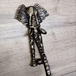 Puede incluir: Un gancho de elefante de metal decorativo con un acabado texturizado. El gancho tiene la forma de un elefante con la trompa enroscada. El gancho está montado en una superficie de madera.