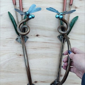 May include: Two handcrafted metal door handles with dragonfly and cattail designs. The handles have a bronze finish with a wood grain pattern. Each handle has a dragonfly with blue wings and green eyes, and cattails with copper-coloured tips.