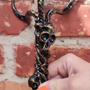 Op de afbeelding: Een gedetailleerde, donkerbronzen sculptuur van een schedelvormig object met gebogen hoorns. De sculptuur heeft een gedraaid, organisch ontwerp. De achtergrond is een bakstenen muur.