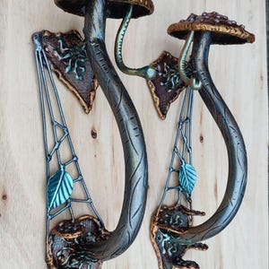 以下が含まれることがあります： スタイリッシュなキノコを模した2つの装飾的な壁掛け燭台。燭台は、赤のアクセントが付いたブロンズ色の茎とキャップを備えています。デザインには、ティールとブロンズの色合いの葉とウェブのようなディテールが含まれています。