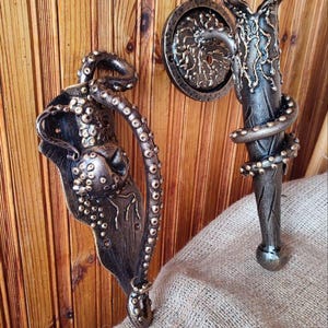 May include: Two ornate, bronze-colored door handles with an octopus theme. One handle features a stylized octopus design, while the other has a tentacle wrapped around a handle. The handles are mounted on a wooden door.