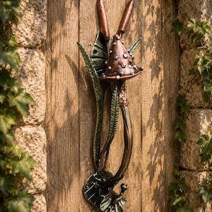 May include: A metal door handle shaped like a mushroom with a copper-colored cap featuring raised dots, a curved stem, and dark green leaves and cattails extending upwards and downwards along the vertical mounting plate on a wooden door.