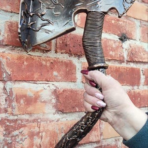 Puede incluir: Hacha de color bronce con detalles intrincados, incluyendo un mango y una hoja texturizados. El hacha se muestra contra una pared de ladrillo rojo, resaltando su diseño rústico y artesanía. La cabeza del hacha tiene patrones decorativos.
