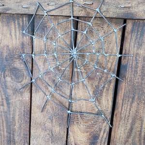 May include: A spider web sculpture constructed from dark grey metal rods joined at intersecting points. The web features a central hub with radiating spokes connected by concentric circular segments, creating a geometric pattern of open spaces.