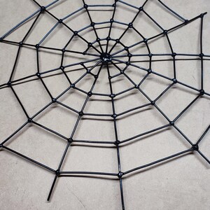 May include: A black spider web decoration constructed from thin, rigid material. The design features a central hub with radiating spokes connected by concentric circular segments, forming a geometric, multi-layered web pattern with pointed ends.
