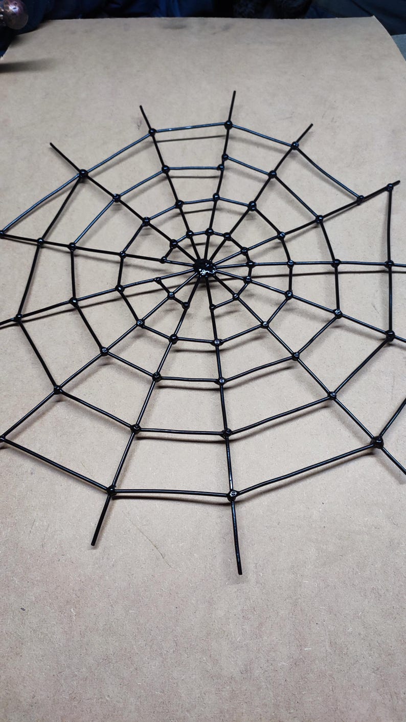 May include: A black spider web decoration constructed from thin, rigid material. The design features a central hub with radiating spokes connected by concentric circular segments, forming a geometric, multi-layered web pattern with pointed ends.