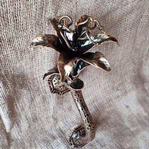 Puede incluir: Una escultura de flor de metal de color bronce con pétalos detallados y un tallo texturizado. La escultura tiene un mango curvo y está sobre un fondo de tela neutra. El centro de la flor es oscuro, contrastando con los pétalos.