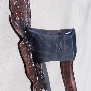May include: A rustic metal wall hook in the shape of an axe with a brown, textured handle and a black metal axe head. The hook is mounted on a wall.