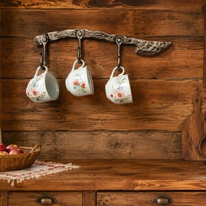 May include: A dark metal wall-mounted rack with three hooks, each holding a white ceramic mug decorated with a colourful floral pattern. The rack features a textured, hammered finish and is attached to a horizontal wooden plank wall surface.