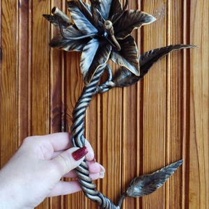 Puede incluir: Escultura de flor de metal hecha a mano con hojas y pétalos detallados. El tallo está torcido y el color general es bronce. La escultura está diseñada para ser montada en la pared.