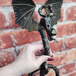 Op de afbeelding: Een bronzen deurklopper in de vorm van een draak. De draak heeft gedetailleerde vleugels, een lange nek en een ringvormige handgreep. De klopper wordt tegen een rode bakstenen muur gehouden.