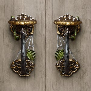 May include: Two matching metal door handles featuring a dark bronze finish with gold highlights. Each handle is shaped like a mushroom with a spotted cap, a central vertical grip, and decorative green leaves and silver web-like accents.