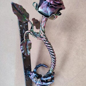 May include: A metal door handle shaped like a rose with a skeletal hand holding the stem. The rose is a deep red color with green leaves. The handle is a dark brown color with a metallic finish.