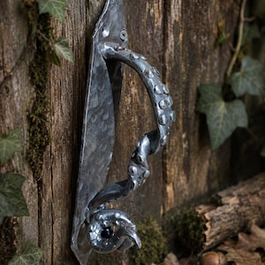 May include: A hand-forged iron door handle featuring a textured, hammered metal backplate and a curved, tentacle-shaped grip adorned with raised circular suction cup details. The dark grey metal has a weathered finish with visible screw heads.