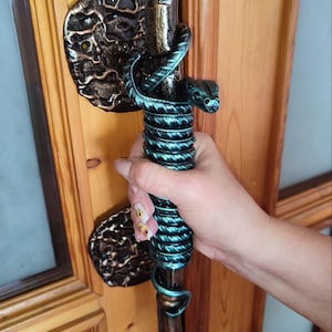 Könnte beinhalten: Verzierter Türgriff mit Bronze-Finish und einer türkisfarbenen Schlange, die sich um den Griff windet. Der Griff ist an einer Holztür mit einer passenden Bronze-Rückplatte befestigt. Die Tür hat Glasscheiben.
