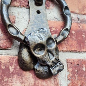 Puede incluir: Aldaba de puerta con forma de calavera de color bronce con un asa de anillo. La calavera está detallada con cuencas de los ojos, una cavidad nasal y dientes. El aldaba está montada en una pared de ladrillos rojos.