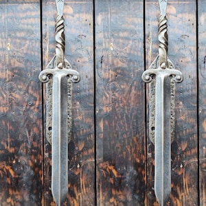 Pode incluir: Dois ganchos decorativos em forma de espada montados em uma parede de madeira. Os ganchos são de metal e têm um design retorcido. Os ganchos são de cor prata escura.