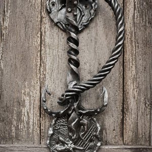 Könnte beinhalten: Ein handgeschmiedeter Türklopfer aus Eisen mit einem gedrehten, seilartigen Griff und einem zentralen Ankermotiv. Der Beschlag ist auf zwei runden Platten mit strukturierter, gehämmerter Oberfläche montiert und in dunklem Metallic-Grau gehalten.
