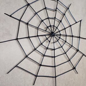 May include: A black spider web decoration constructed from thin, rigid rods arranged in a radial pattern with concentric circular connectors. The structure features a central hub with multiple spokes extending outward to form a geometric web design.