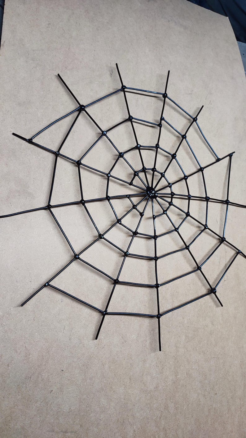 May include: A black spider web decoration constructed from thin, rigid rods arranged in a radial pattern with concentric circular connectors. The structure features a central hub with multiple spokes extending outward to form a geometric web design.