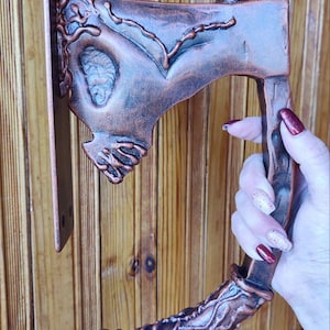 Op de afbeelding: Een bronzen decoratieve deurklink in de vorm van een bijl. De handgreep heeft ingewikkelde reliëfsnijwerk en is gemonteerd op een rechthoekige metalen plaat. De bijlkop heeft een gedetailleerd ontwerp.
