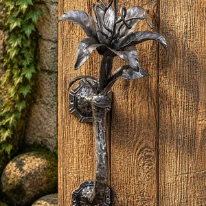 May include: A dark gray, hand-forged metal door handle featuring a sculpted lily flower at the top. The handle has a textured, hammered finish and is secured to a wooden door with two octagonal, bolted mounting plates at the top and bottom.