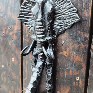 Puede incluir: Un aldaba de puerta de metal negro con forma de elefante. Está fijada a una puerta de madera oscura. El elefante tiene detalles, incluyendo grandes orejas y colmillos. El mango tiene una textura y está fijado con tornillos.
