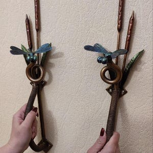 Puede incluir: Dos perchas de pared de color bronce con diseños de libélulas y espadañas. Cada percha presenta una libélula con alas azules y un cuerpo enrollado, junto con espadañas y hojas verdes. Las perchas son sostenidas por manos.