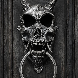 May include: A silver-toned metal door knocker shaped like a horned skull with intricate textured details, deep eye sockets, and prominent fangs. A large, smooth circular ring hangs from the jaw of the skull, serving as the primary striking element.