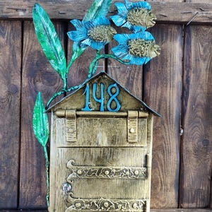 May include: A decorative mailbox with a gold finish, featuring blue floral and fish accents. The mailbox has the number "148" in blue on the roof. The mailbox is mounted on a wooden fence.