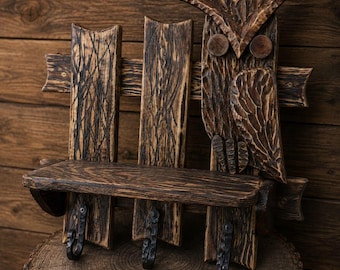 May include: A wall-mounted wooden shelf featuring a carved owl figure on the right side. The unit is constructed from dark, scorched wood planks and includes three black metal hooks attached to the bottom for hanging items.