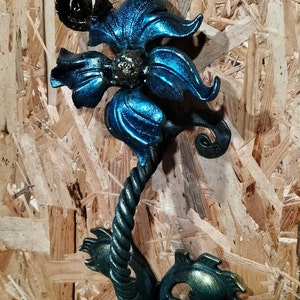 Puede incluir: Escultura floral decorativa de metal con acabado metálico azul y verde. La escultura presenta un diseño floral detallado con un tallo retorcido y formas intrincadas de pétalos. La pieza está montada sobre una superficie de madera.