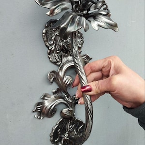 Puede incluir: Manija de puerta plateada y ornamentada con un diseño floral. La manija presenta una flor grande y estilizada en la parte superior, con intrincados detalles de hojas y vides. La manija es sostenida por una mano con esmalte de uñas rojo, montada sobre una superficie gris.