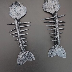 Può includere: Due ornamenti grigi stilizzati a forma di scheletro di pesce. Ogni ornamento presenta un design dettagliato della testa, della spina dorsale e della pinna caudale. Gli ornamenti sono realizzati in un materiale strutturato e sono su sfondo grigio scuro.