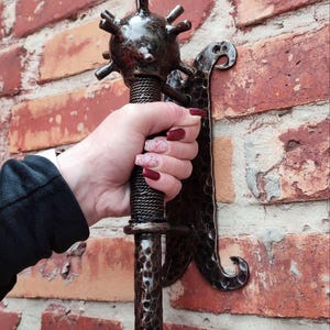 Op de afbeelding: Een donkere, metalen knots met een stekelige kop en een getextureerde handgreep, gemonteerd op een decoratieve beugel. De knots wordt tegen een rode bakstenen muur gehouden.