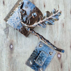 May include: Hand-forged metal cabinet hardware set. The set includes a hammered metal handle with an oak leaf and acorn design, and a matching latch plate. The hardware has a rustic, antique finish.