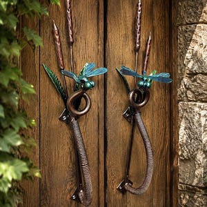 May include: A pair of metal door handles featuring a dragonfly design with blue wings and green bodies. The handles are accented with copper-toned cattail stalks and leaves, and each handle has a coiled, textured grip attached to the door.