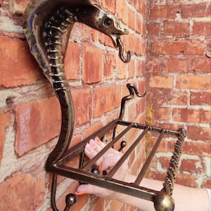 May include: A handcrafted metal wall shelf featuring a cobra design. The shelf is made of dark bronze-colored metal and includes a shelf and two hooks. The cobra's head is at the top, with its hood flared.
