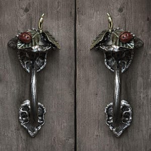 May include: A pair of silver-toned metal door handles featuring a sculpted mushroom design. Each handle is topped with a small red ladybug resting on green leaves, with a curved stem handle and a textured, organic-shaped base plate.