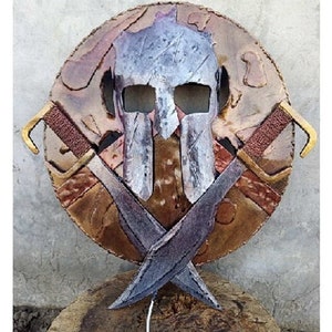 May include: A metal Spartan helmet with a weathered finish, mounted on a circular shield with a copper and gold design. Two crossed swords are attached to the shield.