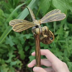 May include: A metallic bronze-colored dragonfly-shaped door handle or wall hook. The dragonfly features textured wings and a long, slender body that extends downward into a curved handle. A circular mounting plate with screw holes is visible.