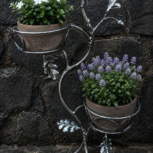 May include: A silver-colored metal plant stand featuring a branching design with decorative oak leaves and acorns. The stand has two circular holders, each containing a brown terracotta pot. One pot holds white flowers, and the other holds purple flowers.