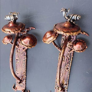 Op de afbeelding: Twee bronskleurige paddenstoelvormige deurgrepen. Elke handgreep heeft een gedetailleerde paddenstoelkap, steel en kleinere paddenstoelaccenten. De handgrepen zijn gemonteerd op een decoratieve, getextureerde achterplaat met een donkergrijze achtergrond.