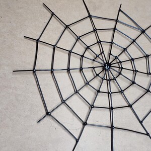 May include: A black spider web decoration constructed from thin, rigid plastic rods arranged in a radial pattern with concentric circles connected by small circular joints, forming a geometric web structure with multiple radiating spokes.