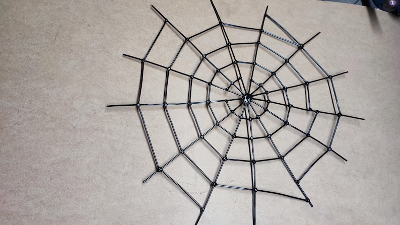 May include: A black spider web decoration constructed from thin, rigid plastic rods arranged in a radial pattern with concentric circles connected by small circular joints, forming a geometric web structure with multiple radiating spokes.