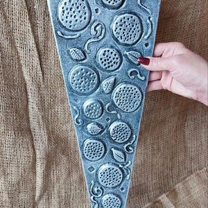 May include: A grey, pizza-shaped wall hanging with raised, textured details. The design includes circular pepperoni and decorative swirls. The piece is held by a hand with a red painted nail. The background is a burlap-like fabric.