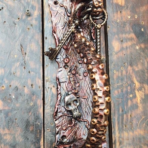 Peut inclure: Une poignée de porte décorative en métal, en forme de pieuvre avec un motif de crâne et d'os croisés. La poignée est peinte dans des tons de brun, rouge et or.